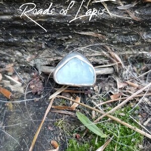 May include: A silver ring with a light blue gemstone set in a triangular shape. The ring is resting on a piece of wood with moss growing on it.