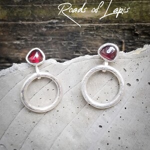 May include: A pair of silver hoop earrings with a red gemstone in the center of each hoop. The hoops are textured and have a hammered finish.