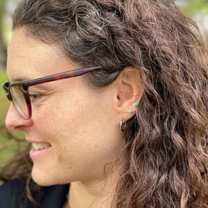 May include: A woman with long, wavy brown hair wearing a pair of small turquoise stud earrings and a silver hoop earring in her left ear.