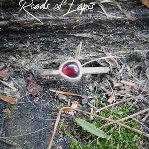 May include: A silver ring with a red gemstone set in a triangular shape. The ring is on a wooden surface with moss and twigs.