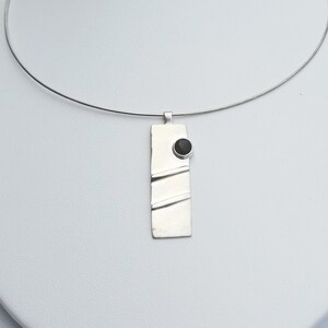 May include: A silver necklace with a rectangular pendant featuring a black stone. The pendant has a textured surface with three horizontal lines.