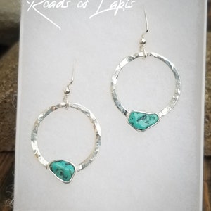 May include: A pair of silver hoop earrings with a turquoise stone accent. The hoops are hammered for a textured look. The earrings are displayed in a white box with the text "Roads of Lapis" written in black.