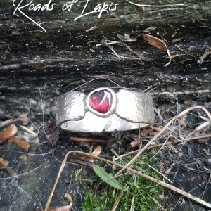 May include: A silver ring with a red gemstone set in the center. The ring is on a wooden surface with moss and twigs.