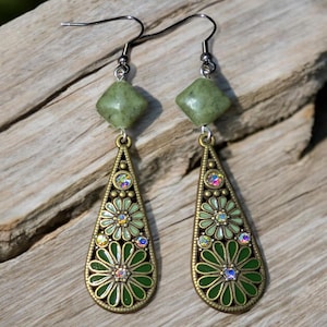 May include: A pair of dangle earrings with a green and gold floral design. Each earring features a green square stone at the top, a teardrop-shaped pendant with green enamel, and small iridescent crystals. The earrings are set against a wooden background.