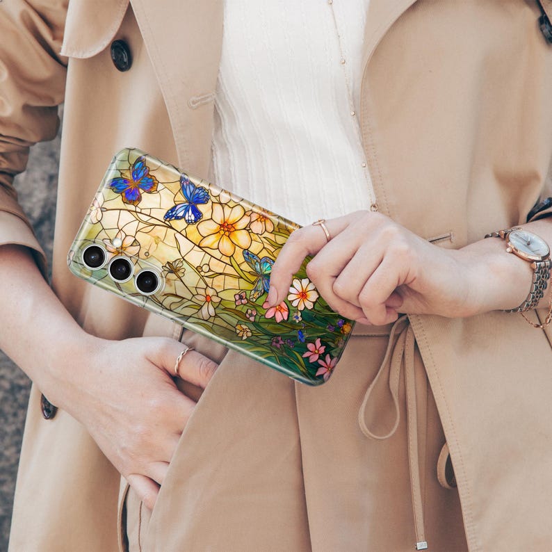 May include: A clear phone case featuring a stained-glass design with blue butterflies, yellow and white flowers, and green foliage. The case has a camera cutout and is held by a person wearing a tan trench coat.