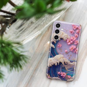May include: A lavender phone case featuring a Japanese wave design with pink cherry blossoms. The case has a three-lens camera cutout and a glossy finish. The wave is depicted in shades of blue and white.