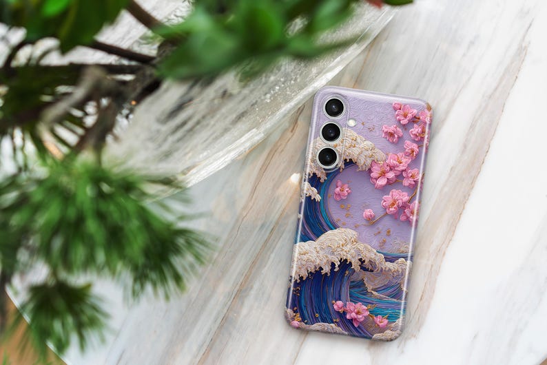 May include: A lavender phone case featuring a Japanese wave design with pink cherry blossoms. The case has a three-lens camera cutout and a glossy finish. The wave is depicted in shades of blue and white.