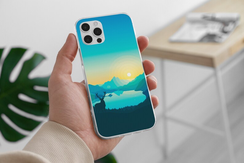 May include: A clear phone case with a blue and green landscape design. The design features a lake, mountains, and a deer silhouette. The phone case is being held in a hand.