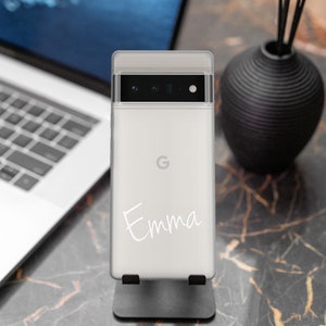Op de afbeelding: Een witte smartphone met een gepersonaliseerde inscriptie die "Emma" luidt. De telefoon heeft een zwarte camerabal met drie lenzen en een Google-logo op de achterkant.