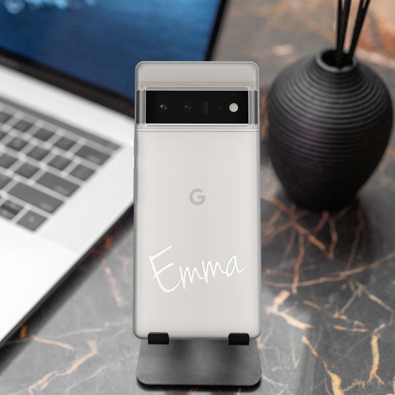Op de afbeelding: Een witte smartphone met een gepersonaliseerde inscriptie die "Emma" luidt. De telefoon heeft een zwarte camerabal met drie lenzen en een Google-logo op de achterkant.