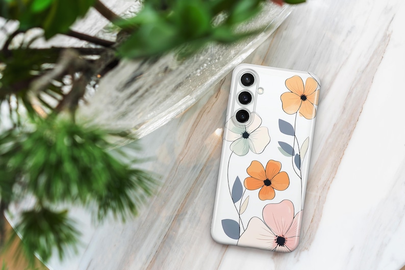 May include: Clear phone case with a floral design. The case features orange, pink, and light green flowers with gray leaves. The phone case has a camera cutout with three lenses.