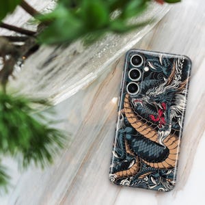 Pu&ograve; includere: Una custodia per telefono nera con un'illustrazione dettagliata di un drago con accenti dorati e rossi. La custodia per telefono ha un ritaglio per l'obiettivo della fotocamera con tre cerchi.