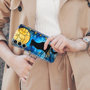 May include: A blue and gold phone case with a black cat silhouette and a moon design. The case has a stained glass look.