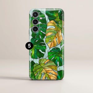 May include: A phone case featuring a stained-glass design of monstera leaves in shades of green and gold. The case has a glossy finish and a light blue background. The phone case has three camera lenses.