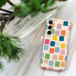May include: A smartphone case with a colorful, abstract design. The case features a pattern of multi-colored squares in shades of pink, orange, yellow, green, and blue on a cream background. The phone case is rectangular and has three camera lenses.