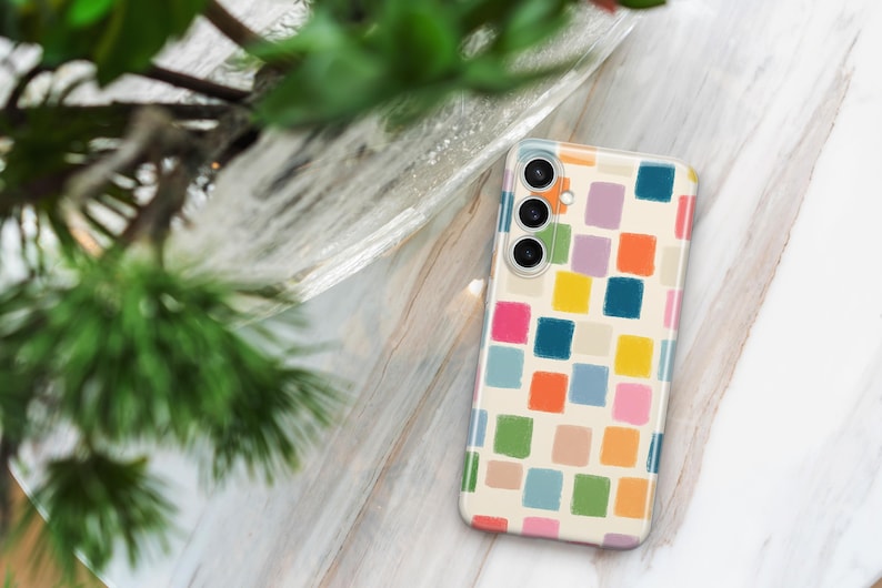 May include: A smartphone case with a colorful, abstract design. The case features a pattern of multi-colored squares in shades of pink, orange, yellow, green, and blue on a cream background. The phone case is rectangular and has three camera lenses.