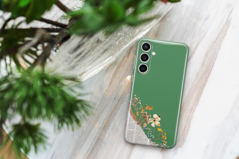 May include: A green smartphone case with a floral design. The case features a collage of dried flowers, butterflies, and newsprint. The phone case is resting on a marble surface, with green foliage in the background.