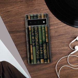 May include: A clear phone case featuring a design of antique books in shades of green, gold, and black. The case is on a wooden surface, with a vinyl record and white earbuds in the background. The phone's camera is visible.