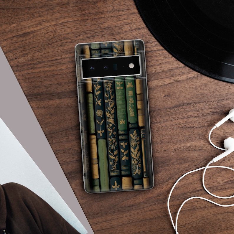 May include: A clear phone case featuring a design of antique books in shades of green, gold, and black. The case is on a wooden surface, with a vinyl record and white earbuds in the background. The phone's camera is visible.