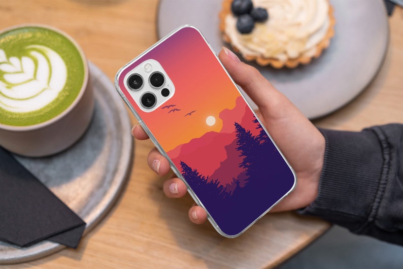 May include: A clear phone case with a sunset landscape design. The case features a silhouette of mountains, trees, and a setting sun. The case is designed for an iPhone with a triple camera lens.