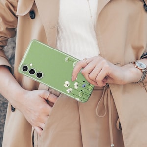 May include: A light green phone case featuring a floral design with white and blue flowers. The case has a camera cutout and is held by a person wearing a tan trench coat and a gold watch.
