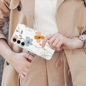 May include: Clear phone case with a floral design featuring blue, orange, and pink flowers with green leaves and black outlines. The case is held against a beige trench coat.