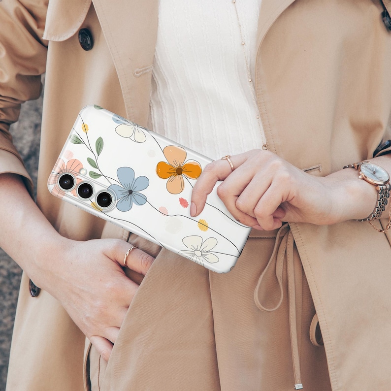 May include: Clear phone case with a floral design featuring blue, orange, and pink flowers with green leaves and black outlines. The case is held against a beige trench coat.