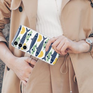 May include: Clear phone case with a repeating pattern of blue fish, yellow lemon slices, and green olive branches on a white background. The case is held in front of a beige trench coat.