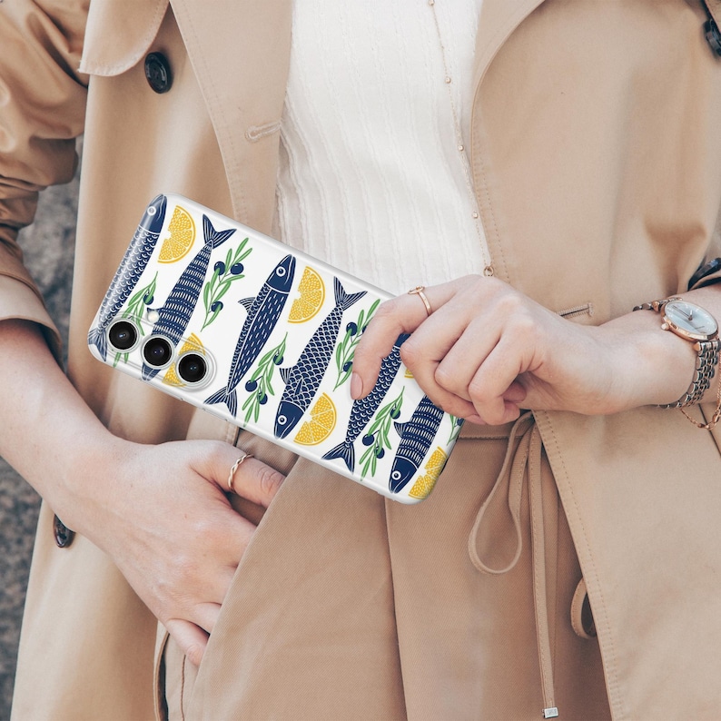 May include: Clear phone case with a repeating pattern of blue fish, yellow lemon slices, and green olive branches on a white background. The case is held in front of a beige trench coat.