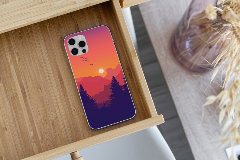 May include: A phone case with a sunset landscape design. The case features a silhouette of mountains, trees, and a bright orange sun. The case is made of a durable material and is designed to protect your phone from scratches and bumps.