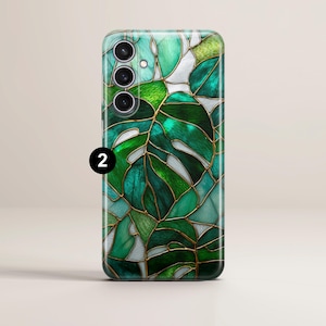 May include: A smartphone case featuring a stained-glass design of a monstera leaf. The case has a glossy finish with shades of green, teal, and white, outlined with gold-colored lines. The phone case has three camera lenses and a flash.