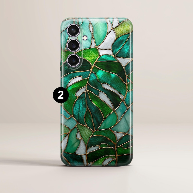 May include: A smartphone case featuring a stained-glass design of a monstera leaf. The case has a glossy finish with shades of green, teal, and white, outlined with gold-colored lines. The phone case has three camera lenses and a flash.