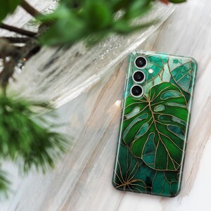 May include: A smartphone case with a green and gold leaf design. The case features a large monstera leaf and other tropical foliage. The case has a glossy finish and a camera module with three lenses. The background is a marble surface.