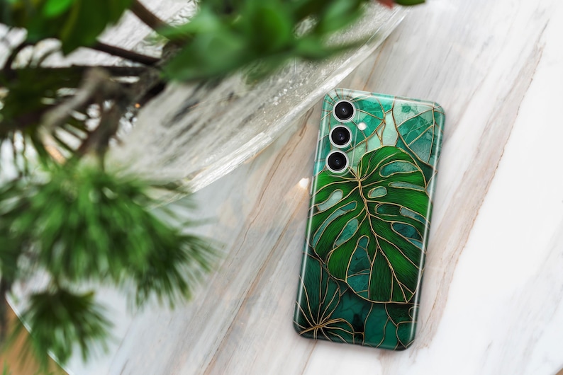 May include: A smartphone case with a green and gold leaf design. The case features a large monstera leaf and other tropical foliage. The case has a glossy finish and a camera module with three lenses. The background is a marble surface.