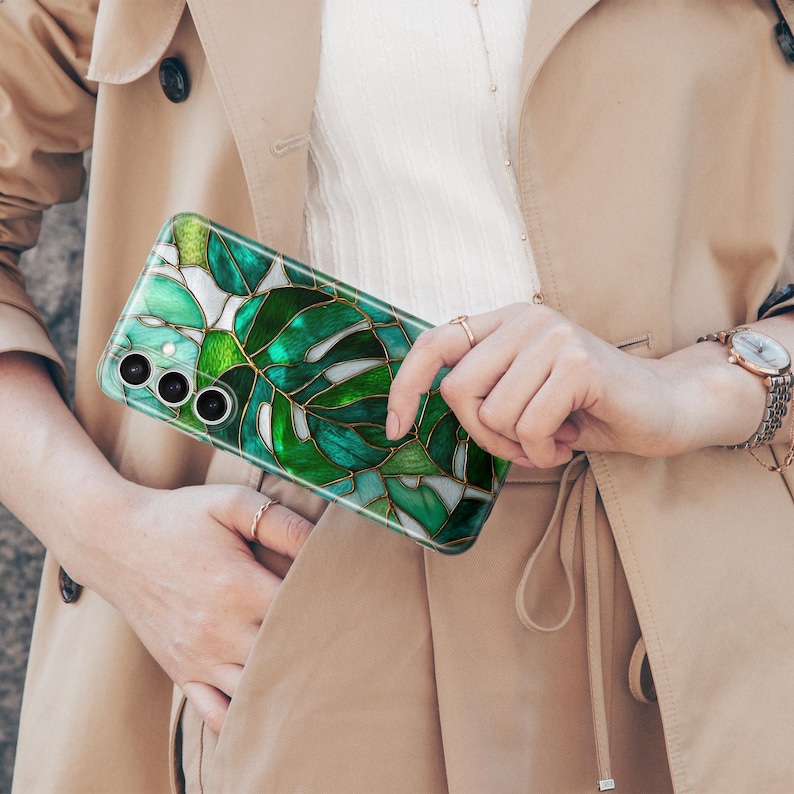 May include: A smartphone case featuring a stained-glass design of green monstera leaves with gold-colored outlines. The case has a glossy finish and a camera cutout. The phone case is held by a person wearing a tan trench coat.