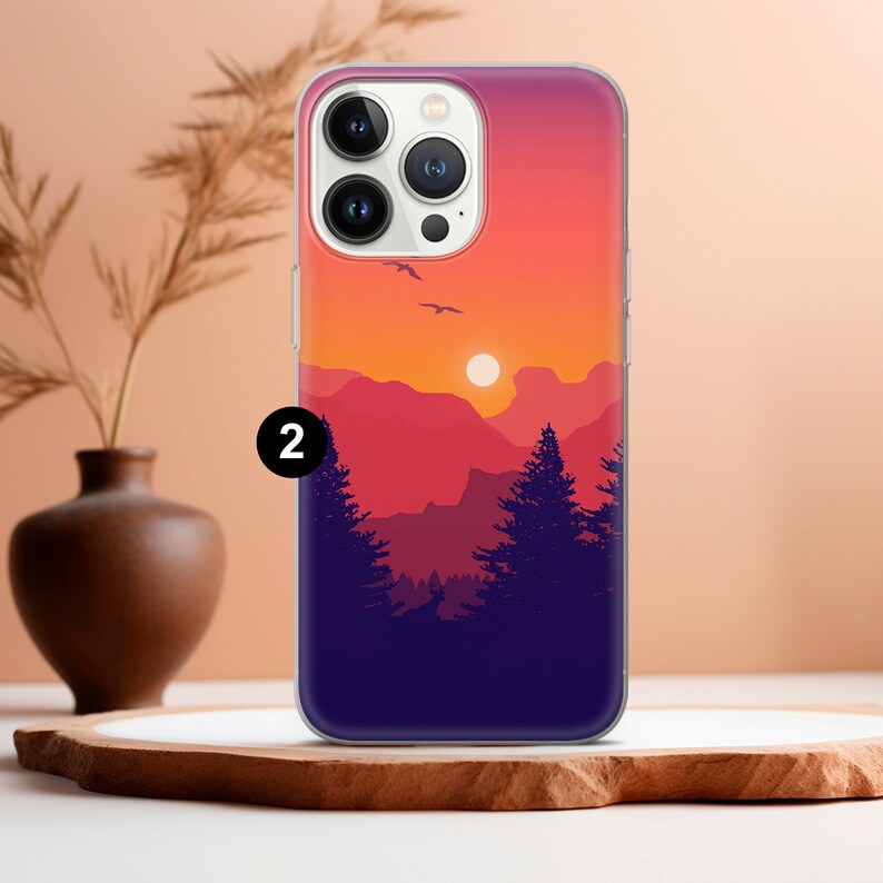 May include: A clear phone case with a sunset scene featuring a mountain range, trees, and a sun. The case has a white border around the camera lens.