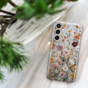 May include: A clear phone case adorned with a floral design. The case features a variety of colorful flowers, including pink, orange, and white blooms. The phone case is lying on a marble surface, with green foliage in the background.