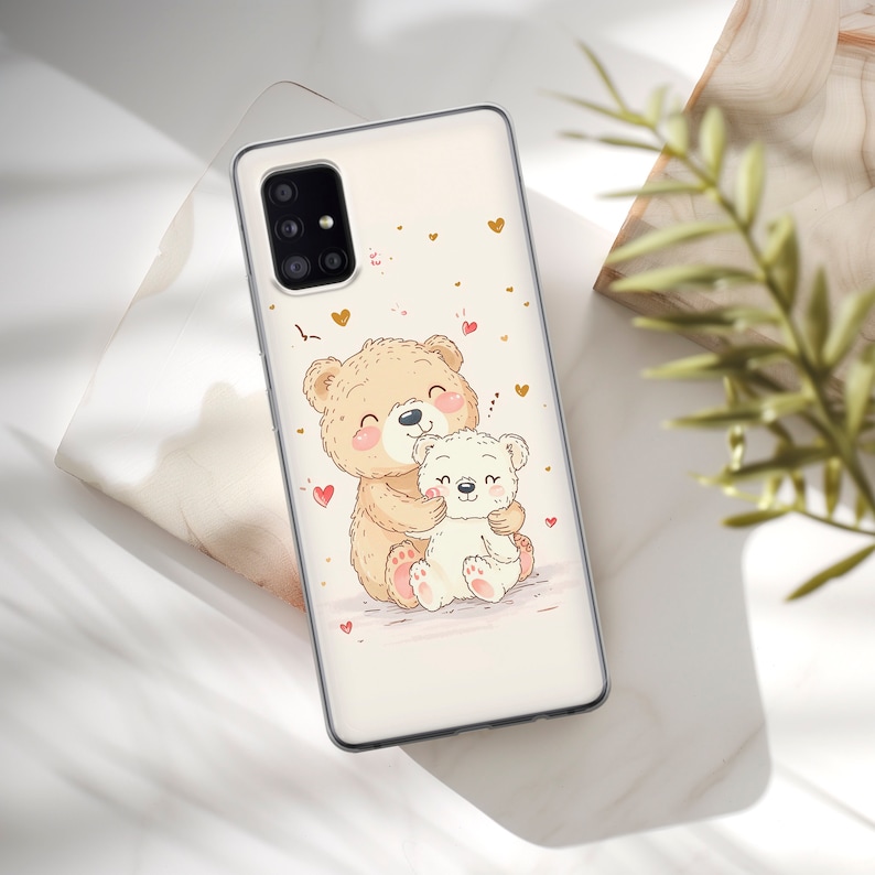 May include: A white phone case with a cute illustration of two teddy bears hugging. The larger bear is brown and the smaller bear is white. The background is white with pink and gold hearts.