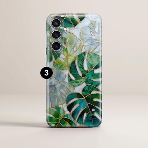 May include: A smartphone case featuring a stained glass design with green monstera leaves and gold outlines. The case has a glossy finish and a camera module with three lenses. The number "3" is printed on the bottom left.