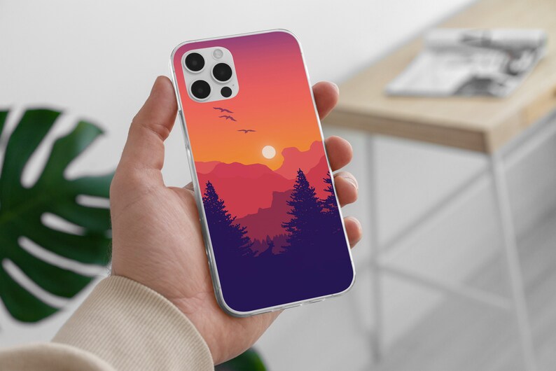 May include: A clear phone case with a sunset landscape design. The design features a silhouette of mountains, trees, and birds flying in a purple, orange, and pink sky.