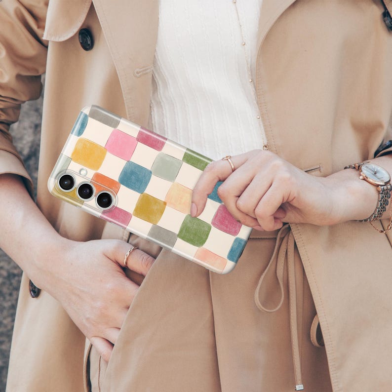 May include: A smartphone case featuring a colorful checkerboard pattern in shades of yellow, orange, pink, blue, green, and gray. The case is held by a person wearing a tan trench coat and a gold watch.