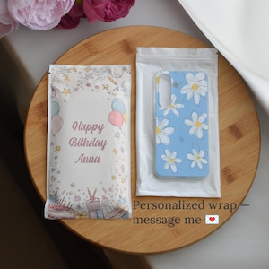 May include: A light blue phone case with white daisy flower designs is displayed in a clear plastic bag. Next to it is a gift bag with the text "Happy Birthday Anna" and illustrations of balloons and a cake.