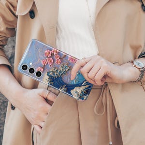 May include: A clear phone case featuring a 3D design of a blue ocean wave with gold accents and pink cherry blossoms. The case is designed for a smartphone with three camera lenses.