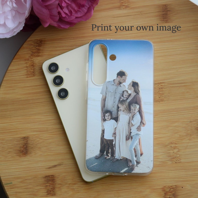 May include: A smartphone case with a family photo print. The case is clear with a blue and white sky design at the top. The phone is a light beige color. The text "Print your own image" is visible.
