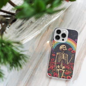 May include: A white iPhone case with a colorful design featuring a skeleton standing in a field of flowers with a rainbow in the background. The case has a camera cutout with three lenses.