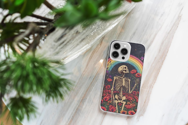 May include: A white iPhone case with a colorful design featuring a skeleton standing in a field of flowers with a rainbow in the background. The case has a camera cutout with three lenses.