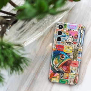 May include: A patterned phone case with a fish illustration and various other images. The case has a collage design with a blue, green, and orange fish. The phone case is on a light-coloured surface, with green foliage in the background.