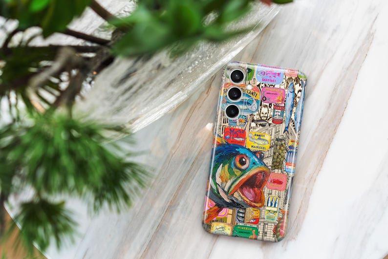 May include: A patterned phone case with a fish illustration and various other images. The case has a collage design with a blue, green, and orange fish. The phone case is on a light-coloured surface, with green foliage in the background.