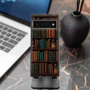 May include: A smartphone case featuring a detailed bookshelf design with rows of books in shades of brown and green. The case is displayed on a black stand, with a laptop and a black vase in the background.