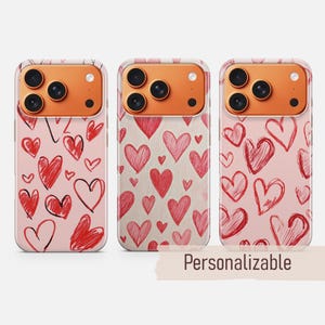 Peut inclure: Trois coques de téléphone avec des motifs de cœurs rouges sur fond rose. Les coques ont une couche protectrice transparente et une bosse pour l'appareil photo. Le texte "Personnalisable" est affiché en bas. Les coques sont conçues pour un smartphone.